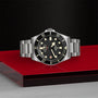 The TUDOR Pelagos LHD, a stainless steel wristwatch with a black dial and bezel, is displayed on a red and silver surface.