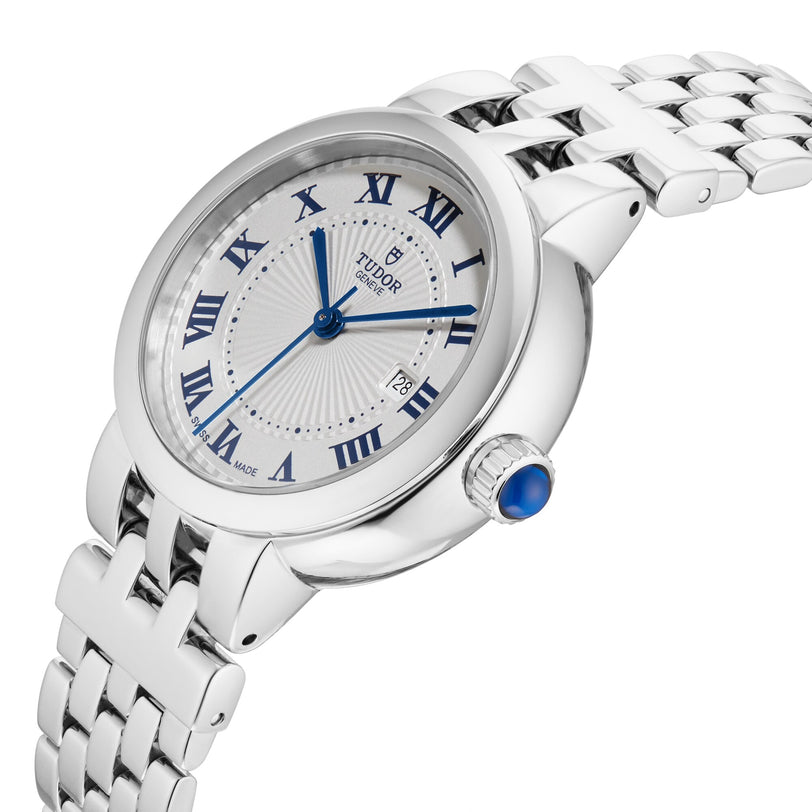 The TUDOR Clair de Rose is a silver wristwatch featuring blue Roman numerals, a date display, and a silver metal link band.