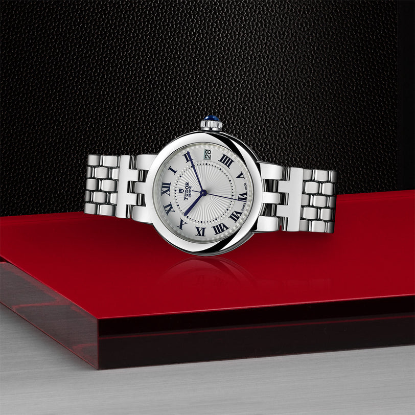 The TUDOR Clair de Rose silver wristwatch with Roman numerals rests on a red and gray surface, set against a black textured background.