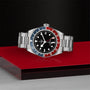 The TUDOR Black Bay GMT is a stainless steel wristwatch with a red and blue bezel, black dial, and metal bracelet displayed on a red surface.