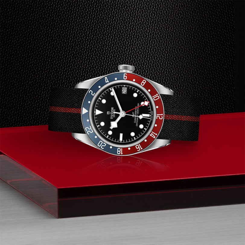A TUDOR Black Bay GMT wristwatch with a black dial, blue and red bezel, and black strap is displayed on a glossy red and black surface.