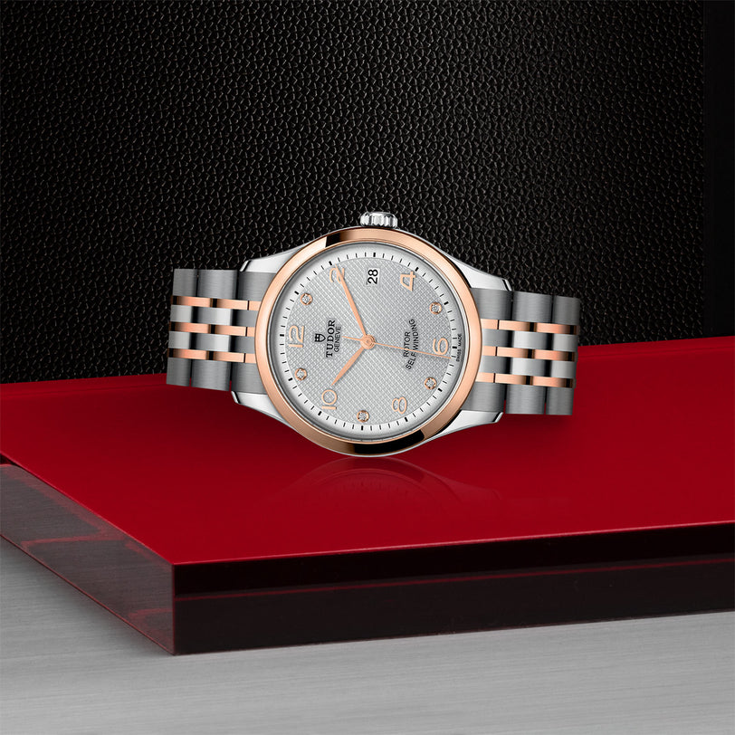 The TUDOR 1926 silver and rose gold wristwatch is showcased on a red and gray surface with a textured black background.
