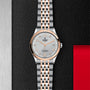 A TUDOR 1926 wristwatch in silver and rose gold features a round face, date display, metal link strap, and sapphire crystal lens, showcased on a textured black and metallic background with a red accent.