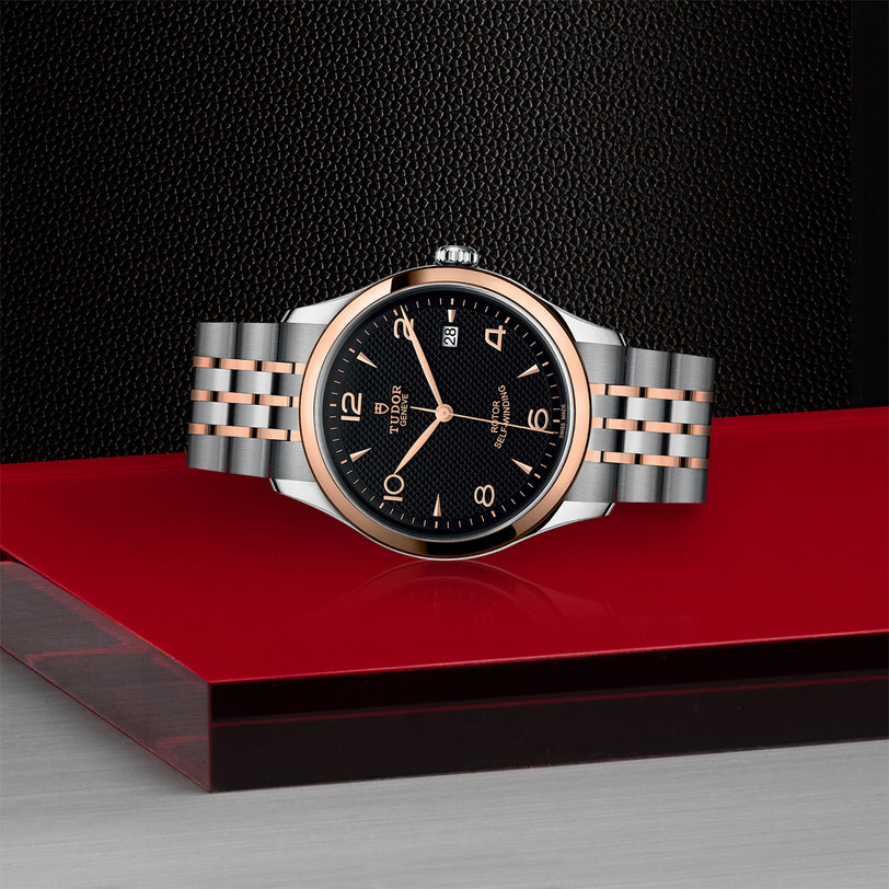 A TUDOR 1926 luxury wristwatch with a black dial, gold bezel, and gold-silver bracelet is elegantly displayed on a red and black geometric surface, highlighting the timeless style of mechanical watches.