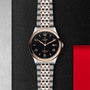 A TUDOR 1926 wristwatch with a black dial, gold hour markers, and a two-tone silver and gold bracelet is displayed on a textured black and silver background with a red accent, highlighting TUDORs heritage.