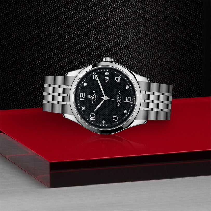 The TUDOR 1926 is a classic stainless steel wristwatch with a black dial, silver hour markers, and a metal bracelet, shown on a red and black platform.