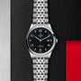 The TUDOR 1926 wristwatch with a black dial and metal bracelet is displayed on a textured black surface.