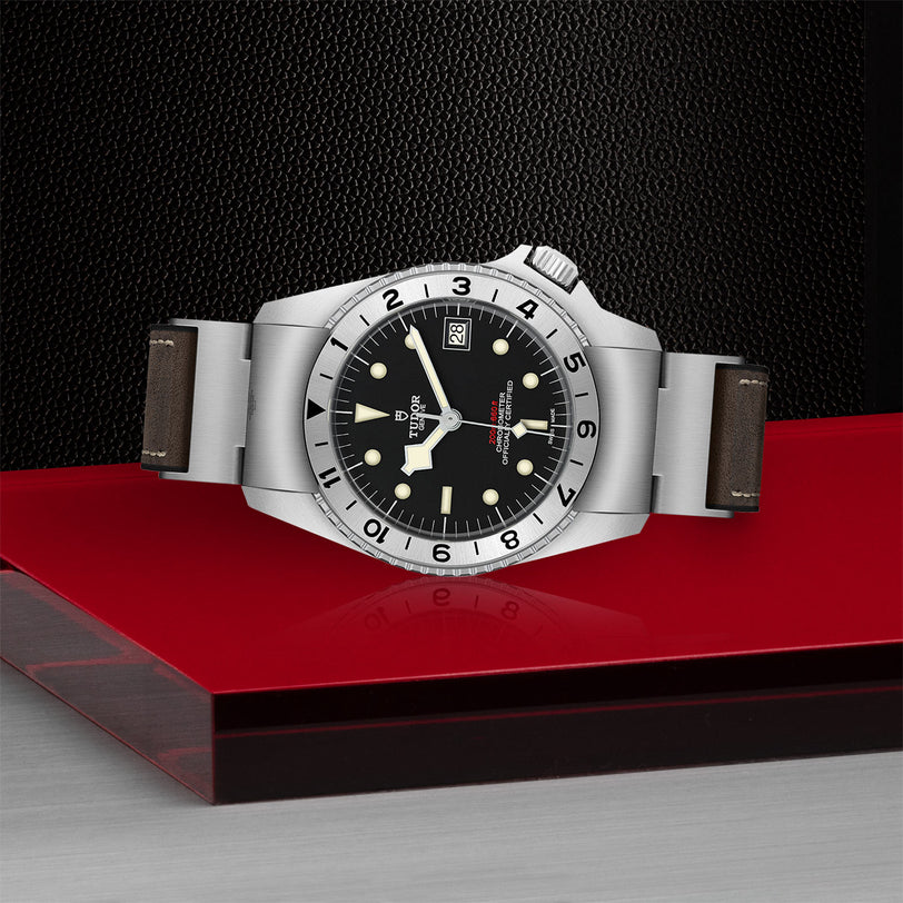 The TUDOR Black Bay P01, a silver wristwatch with a black dial and brown strap, is displayed on red and black surfaces.