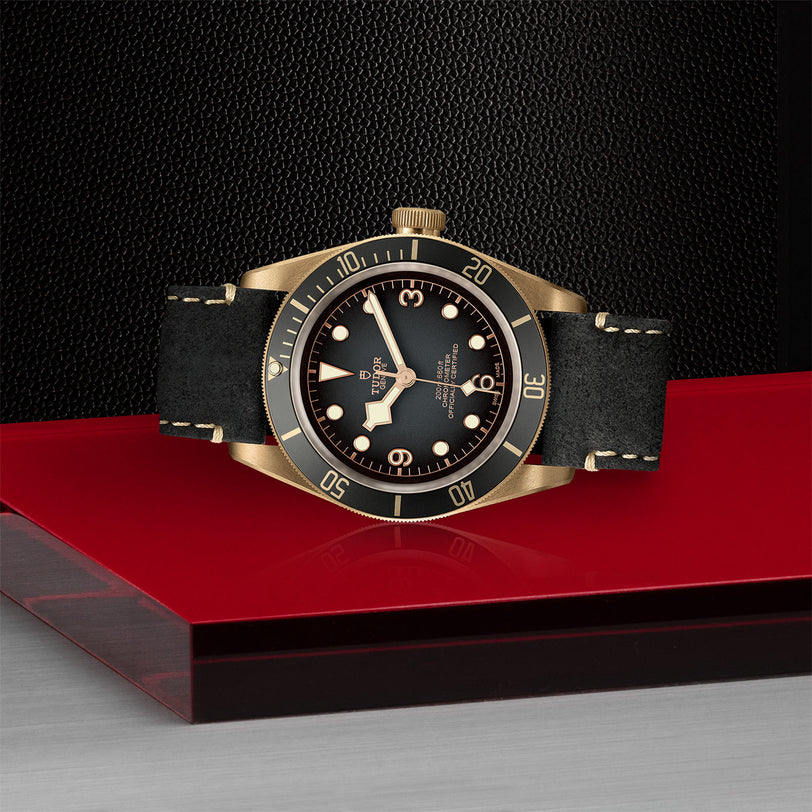 The TUDOR Black Bay Bronze wristwatch, featuring a black dial and strap, is showcased on a red and silver platform against a textured black background.