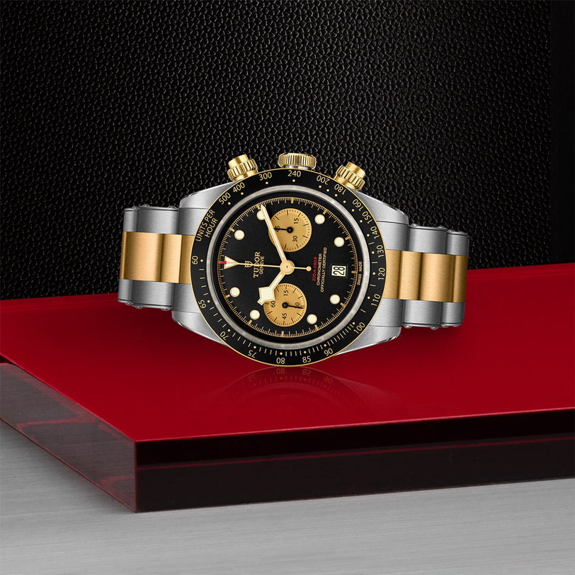 The TUDOR Black Bay Chrono S&G, a two-tone stainless steel and gold watch with a black and gold dial and Swiss-made movement, is displayed on a red and silver surface against a textured black background.