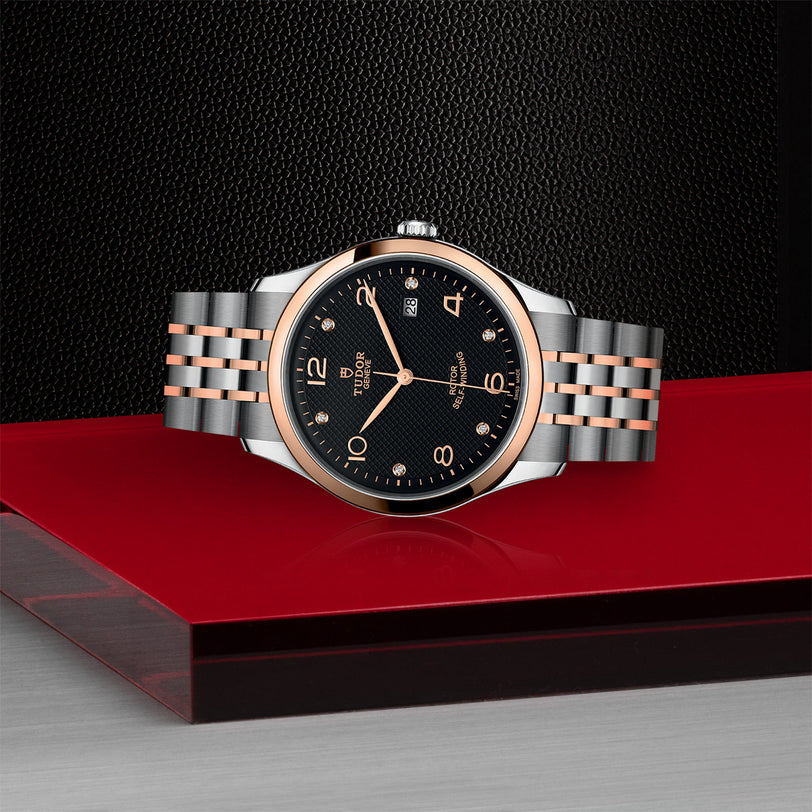 A TUDOR 1926 watch featuring a black dial, rose gold and silver hour markers, and a two-tone metal band rests on a glossy red surface against a textured black backdrop, highlighting the refined style of TUDOR’s mechanical timepieces.