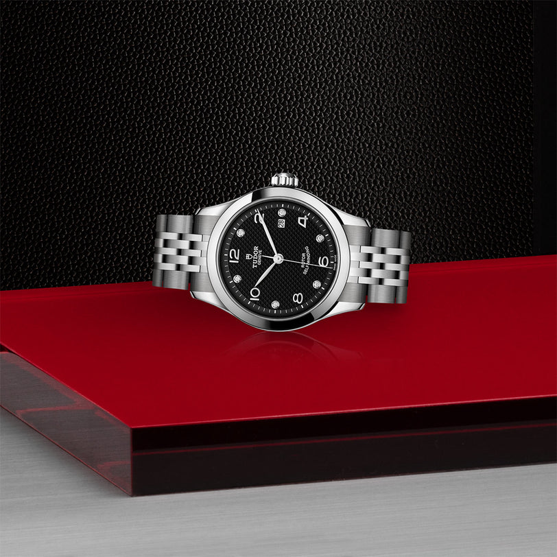 The TUDOR 1926, a timelessly elegant silver wristwatch with a black face and metal band, is showcased on a red and silver platform against a textured black background.