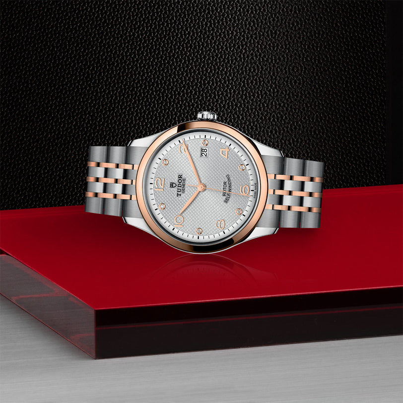 A TUDOR 1926 wristwatch with a silver and rose gold bracelet is displayed on a red and black surface against a textured black background, highlighting the refined elegance of mechanical timepieces.