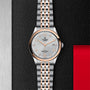 A TUDOR 1926 wristwatch with a silver and rose gold metal band, white textured dial, and date display sits on a textured black surface beside metallic and red panels—a sophisticated option for watch enthusiasts.