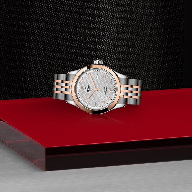 The TUDOR 1926 wristwatch, crafted in silver and rose gold with a textured white dial, is displayed on a glossy red and black surface against a dark background, highlighting its classic mechanical elegance.