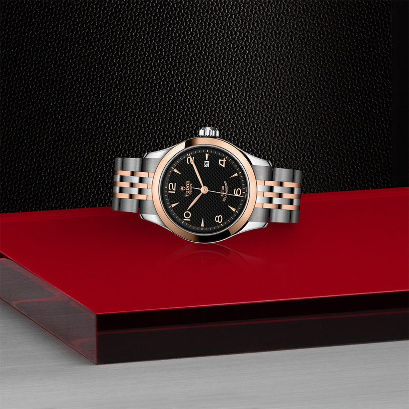 The TUDOR 1926, a classic two-tone wristwatch with a black dial and silver and rose gold bracelet, is displayed on a glossy red and black surface with a textured background, highlighting the elegance of mechanical watches.