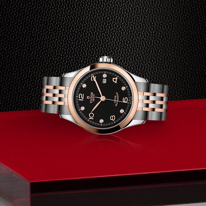 A TUDOR 1926 wristwatch featuring a black textured dial, diamond hour markers, and a two-tone metal bracelet is displayed on a red surface against a textured black background—a modern classic with timeless appeal.
