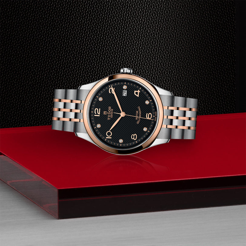 The TUDOR 1926 wristwatch, featuring a black dial, silver and rose gold bracelet, and diamond hour markers, is displayed on a red and black platform with a textured black background.