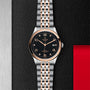 The TUDOR 1926 watch, featuring a black dial, gold accents, and a two-tone metal bracelet, is elegantly displayed on a textured black and steel surface with a red accent.