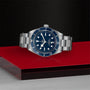 A TUDOR Black Bay 58 watch with a blue dial and bezel is showcased on a red and black geometric surface, emphasizing its iconic design inspired by classic diving watches.