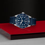 The TUDOR Black Bay 58 wristwatch with a leather strap is displayed on a red and black geometric surface.