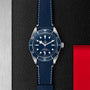 A TUDOR Black Bay 58 wristwatch with a blue dial and leather strap rests on a textured black surface, featuring metal and red accents.