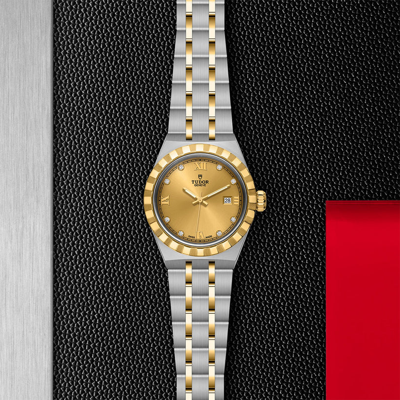 A TUDOR Royal wristwatch with a gold dial, gold bezel, and stainless steel and gold bracelet is displayed vertically on a textured black and silver surface with a red accent.