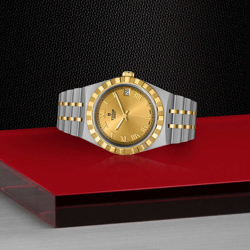 A TUDOR Royal wristwatch with a gold face and Roman numerals is displayed on a red and metallic surface, its stainless steel and gold design contrasting against the black textured background.