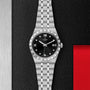 A TUDOR Royal wristwatch featuring a black dial with Roman numerals, date display, and silver bracelet is showcased on a textured black and metallic background.