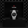 A silver TUDOR Royal wristwatch with a black dial is shown standing in an open black TUDOR box, set against a textured black background that highlights the automatic sport-chic appeal of the Royal collection.
