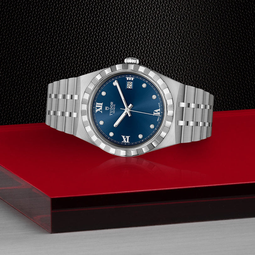 The TUDOR Royal wristwatch features a stainless steel case, blue dial with Roman numerals and date display, and an integrated bracelet, showcased on a glossy red and gray surface against a black textured background.