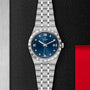 The TUDOR Royal wristwatch features a blue dial with Roman numerals, date display, and an integrated bracelet, shown on a black and metal surface with a red accent—an ideal automatic sport-chic choice for watch enthusiasts.