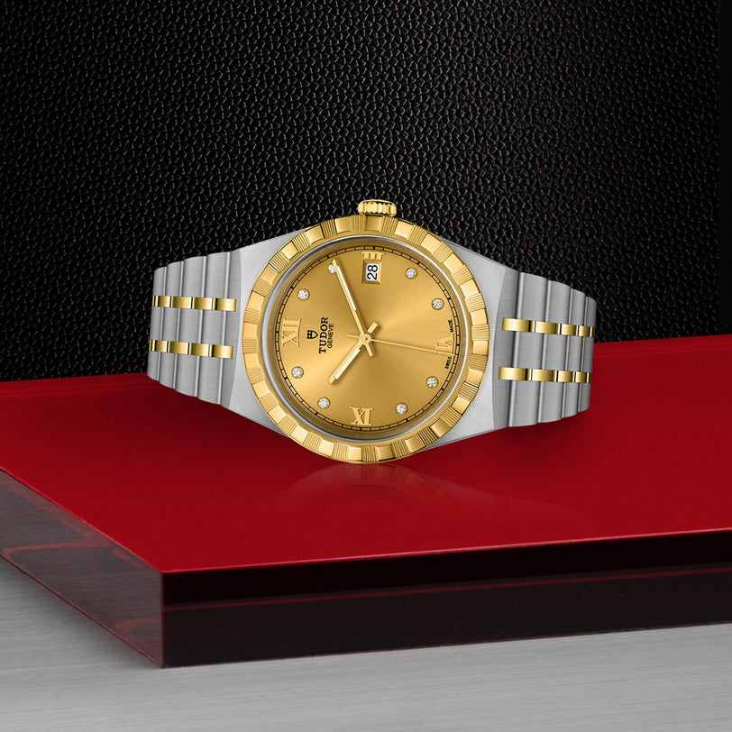 A TUDOR Royal watch with a gold dial, diamond markers, date display, and a stainless steel and gold bracelet is shown on a red and black surface.