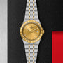 A TUDOR Royal wristwatch with a gold dial, diamond hour markers, and date display in stainless steel and gold is displayed on a textured black and red background.