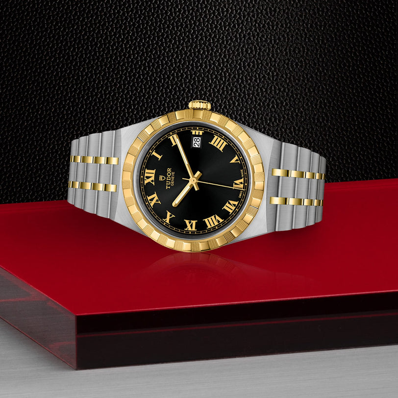 A TUDOR Royal wristwatch with a gold and silver finish, black dial, Roman numerals, date display, and integrated bracelet is showcased on a red and glass surface against a textured black backdrop—exemplifying sport-chic style.