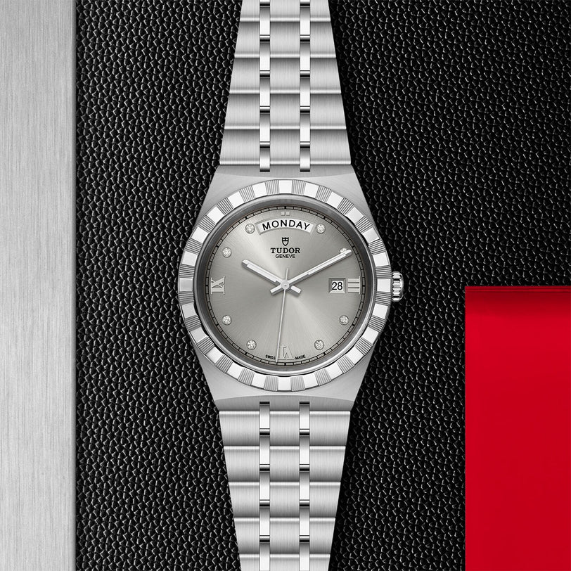 A TUDOR Royal wristwatch with an integrated bracelet, day-date display, and diamond hour markers rests on a textured black surface featuring metallic and red accents.