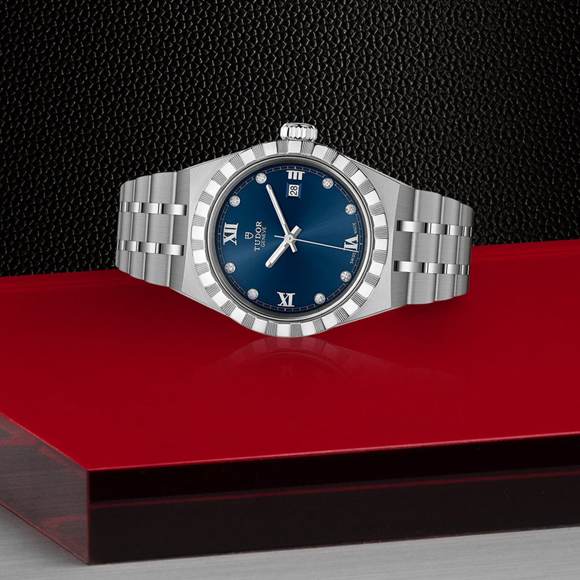 A TUDOR Royal wristwatch featuring a silver case, blue dial, and integrated bracelet is displayed on a red and black surface.