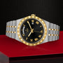 A TUDOR Royal wristwatch with a black dial, gold Roman numerals, and day-date display on an integrated bracelet is shown on a textured red and black surface.