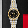 A TUDOR Royal wristwatch with a black dial, gold Roman numerals, day and date display, and a silver-gold bracelet is displayed on a textured black surface.