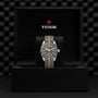 A TUDOR Black Bay 58 925 wristwatch with a fabric strap is displayed upright in an open black TUDOR box, highlighting its iconic 39mm case against a textured background.