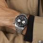 A person wearing the TUDOR Black Bay Chrono, a stainless steel chronograph with black bezel and black-and-white dial, is shown against a beige jacket.