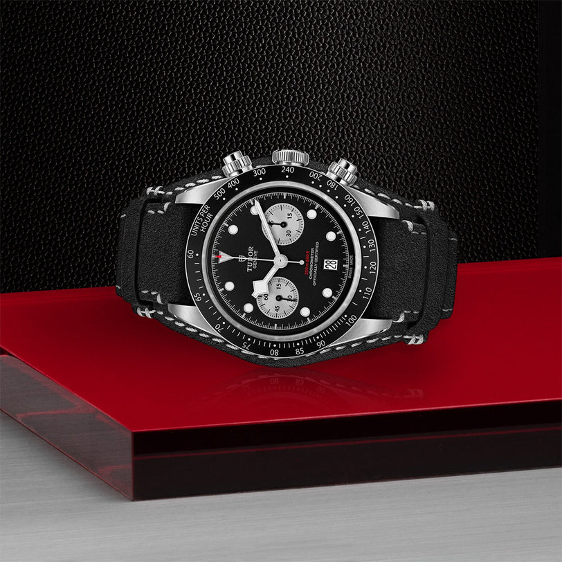 The TUDOR Black Bay Chrono wristwatch, featuring a black strap and a black and silver design, is displayed on a glossy red and gray surface against a textured black background.