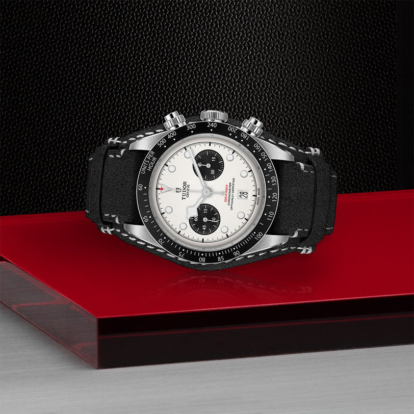 The TUDOR Black Bay Chrono, a stainless steel chronograph with a tachymetric scale, black bezel, white dial, black subdials, and black strap is displayed on a red and gray geometric surface.
