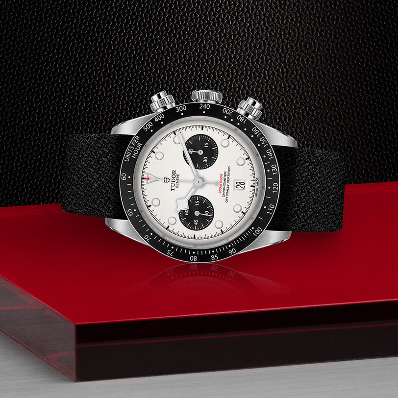 The TUDOR Black Bay Chrono wristwatch, featuring a black fabric strap and a black-and-white dial with two subdials, is shown on a red and black textured surface.