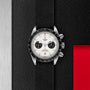 A TUDOR Black Bay Chrono wristwatch with a white dial and black tachymeter bezel sits on a textured black surface next to a metallic panel and red accent.