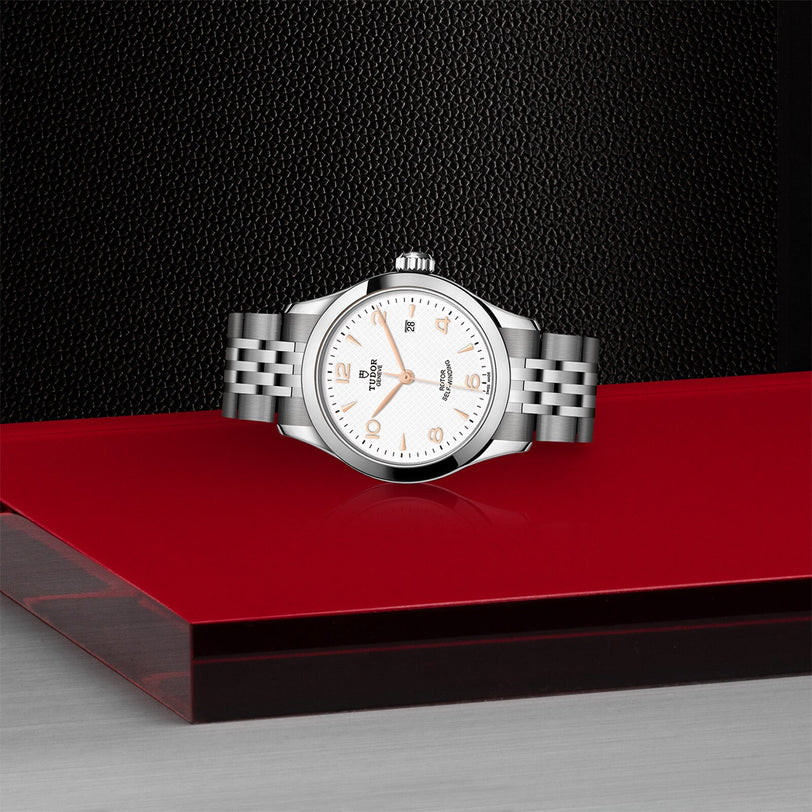 The TUDOR 1926, a silver wristwatch with a white dial and metal bracelet, is showcased on a glossy red and black platform against a textured black background.