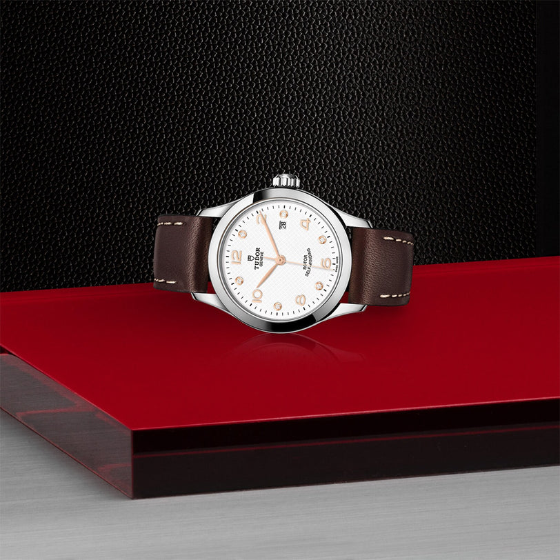 The TUDOR 1926, an elegant watch with a brown leather strap and white dial, is displayed on a red surface against a dark textured background, highlighting its timeless appeal.