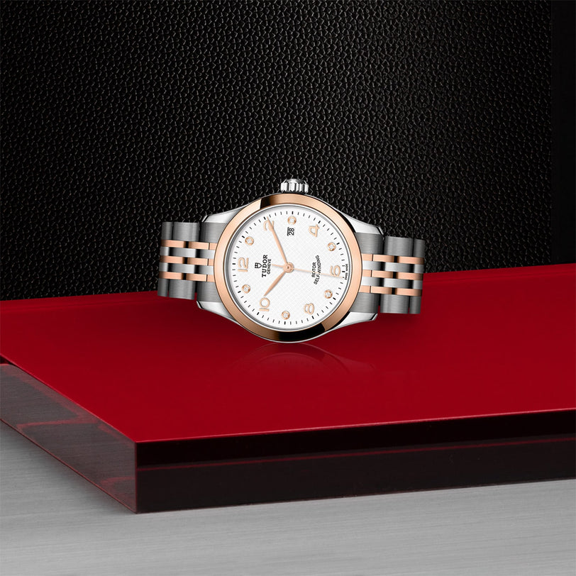 The TUDOR 1926 wristwatch, with a silver and rose gold metal band, rests on a glossy red surface against a textured black background.