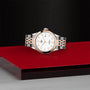 The TUDOR 1926 wristwatch, with a silver and rose gold metal band, rests on a glossy red surface against a textured black background.