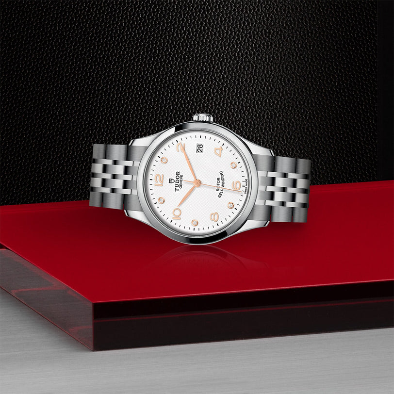 A TUDOR 1926 silver wristwatch with a white dial and metal bracelet is showcased on a red and black platform, set against a dark textured background.